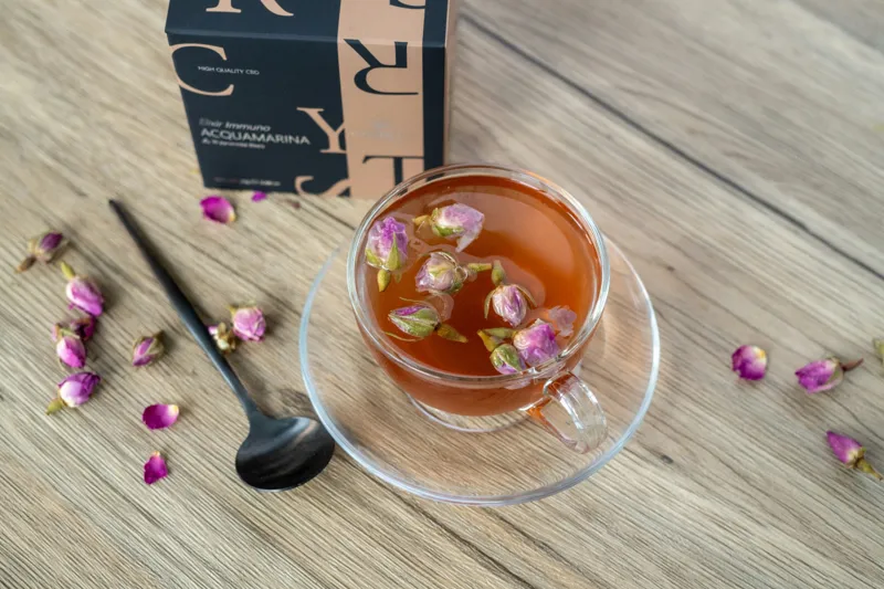 Tisana Crystaltea Acquamarina con fiori di boccioli di rosa in tazza di vetro su tavolo di legno chiaro, accompagnata dalla confezione elegante nera e beige.